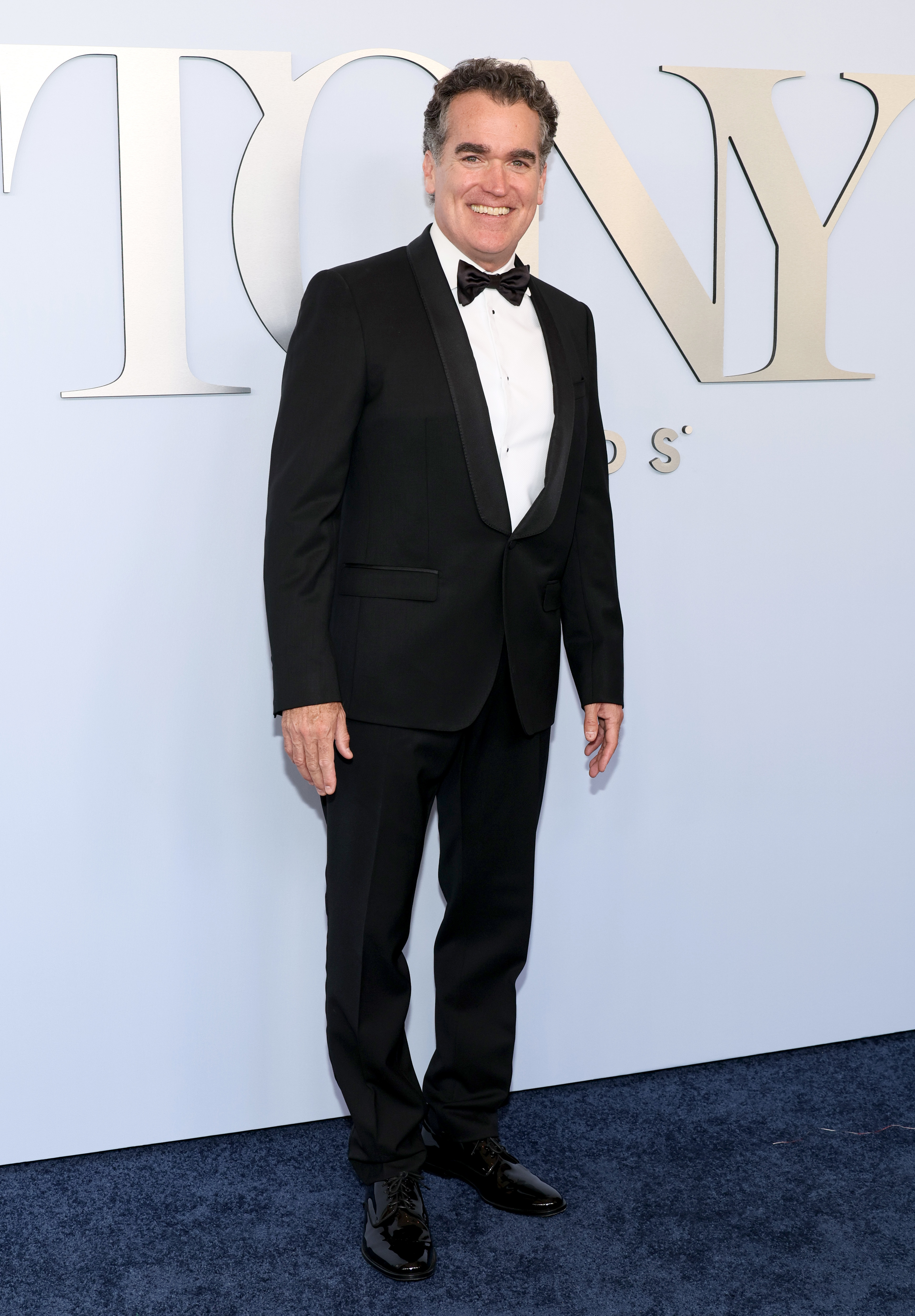 Brian d'Arcy James wearing a black tuxedo with a bow tie, standing on a blue carpet at the Tony Awards