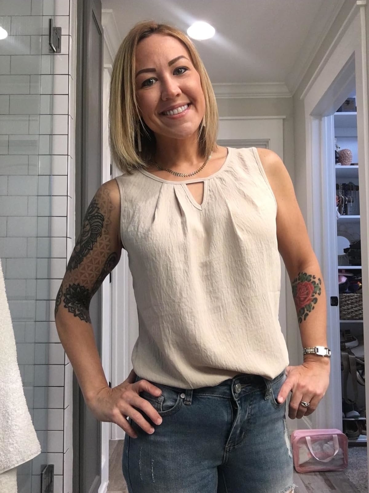 A reviewer with tattoos on both arms smiles while wearing a sleeveless top and jeans, standing in a bathroom