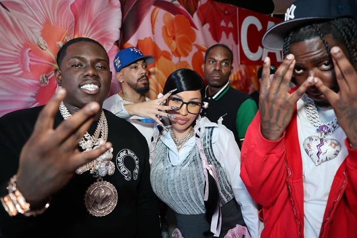 A group of five people poses for the camera on a floral background. The woman in the center wears glasses and a stylish outfit. The men around her wear casual streetwear. Names unknown