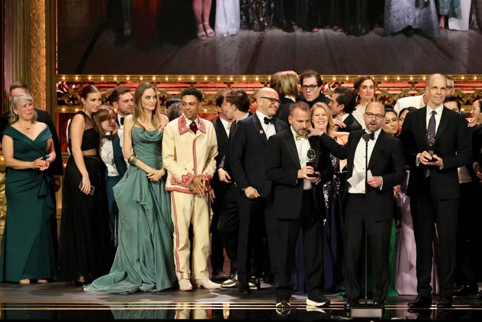Group of actors and creators in formal clothing on stage, holding awards at a ceremony. Multiple people smiling and standing together