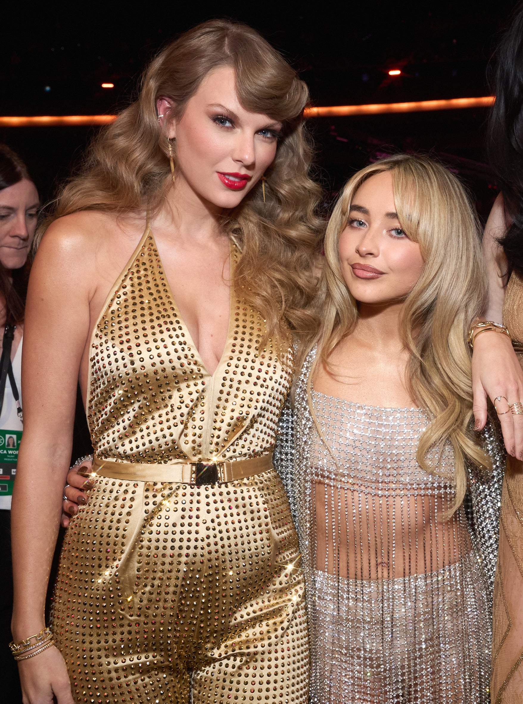 Taylor Swift and Sabrina Carpenter at an event. Taylor wears a metallic jumpsuit, Sabrina wears a sheer glittering dress