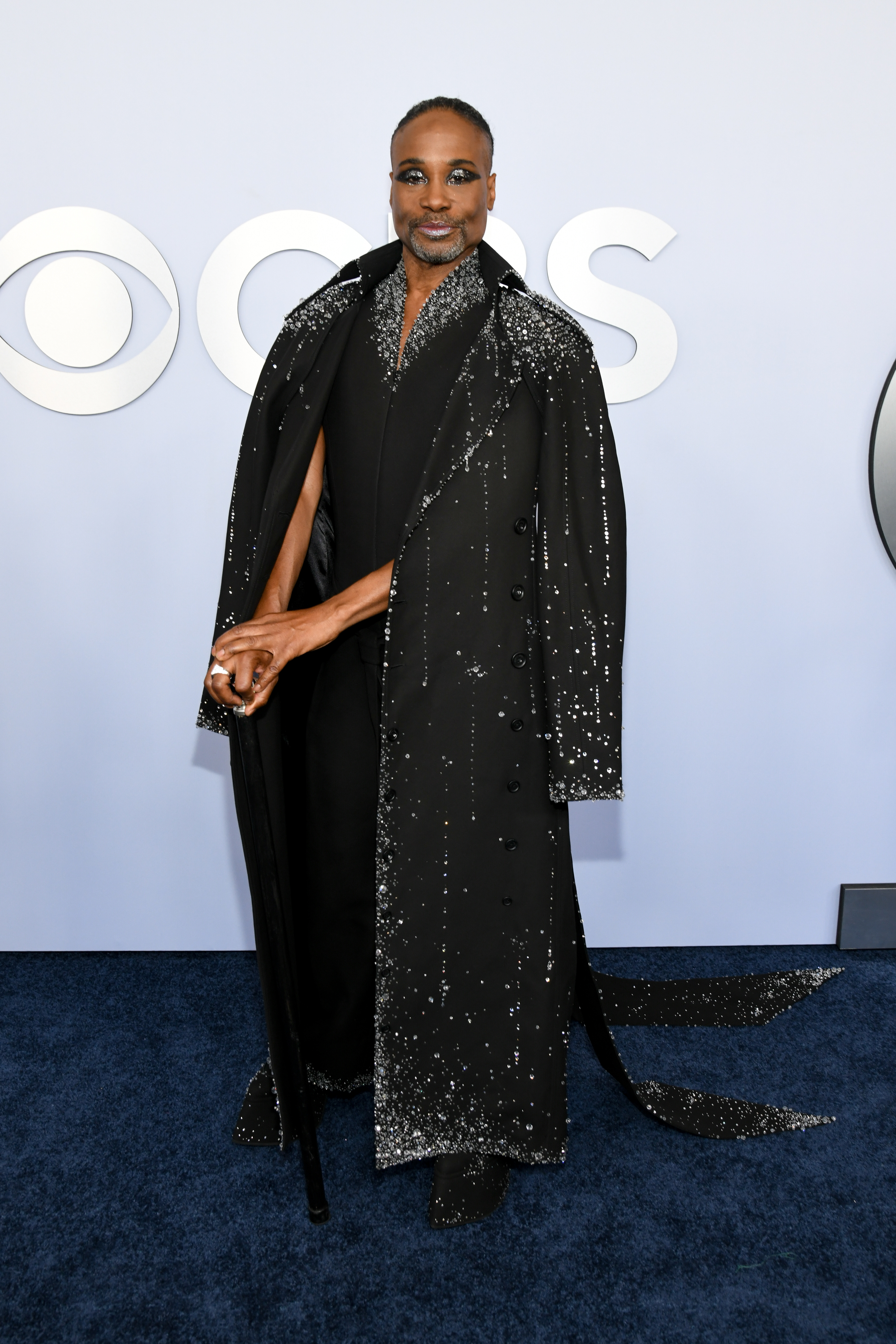 Billy Porter poses on the red carpet, wearing a sparkling black coat over a matching black outfit, holding a cane