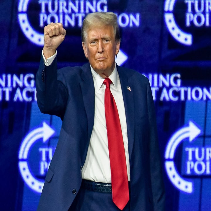 Donald Trump stands in a suit with a red tie, raising his fist at a Turning Point Action event
