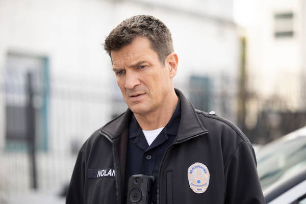 Nathan Fillion, wearing a police uniform with a body camera, looks pensive while on an urban street setup for a scene