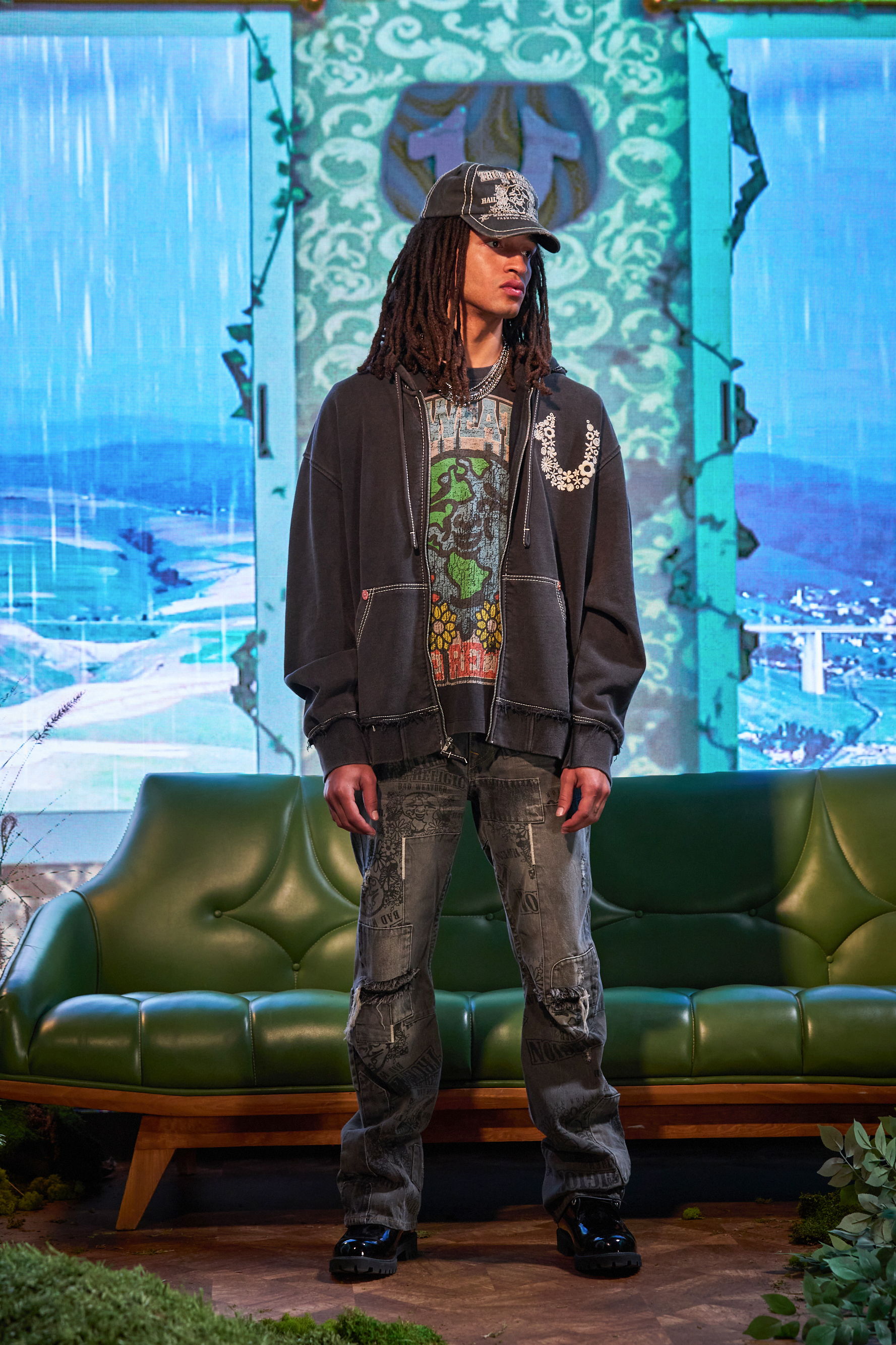Model wearing a patterned baseball cap, graphic shirt, hoodie, and distressed jeans stands in front of a scenic digital backdrop during a fashion show