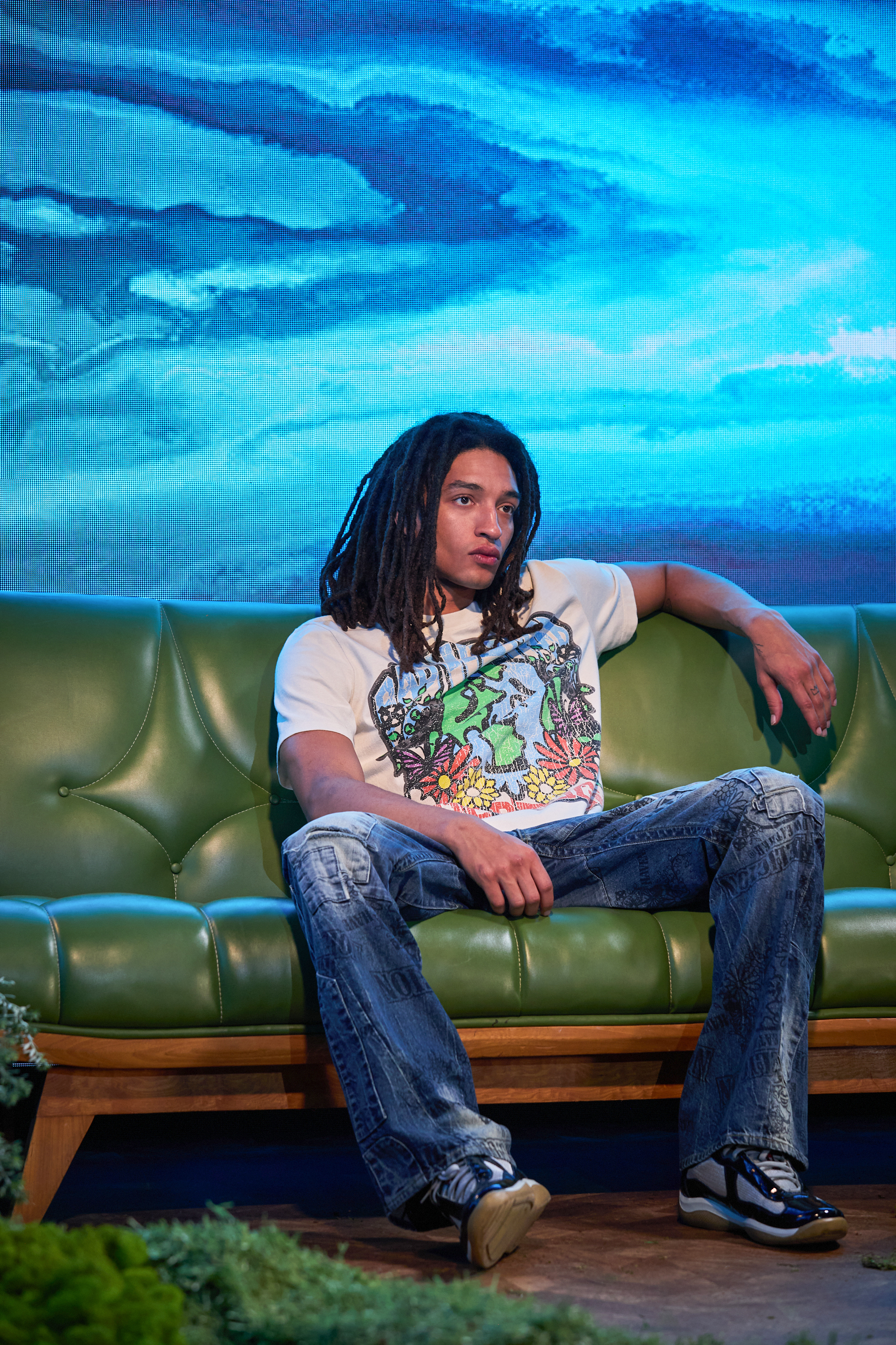 Person sits on green couch, wearing jeans and a graphic t-shirt with a nature design, with a blue scenic background