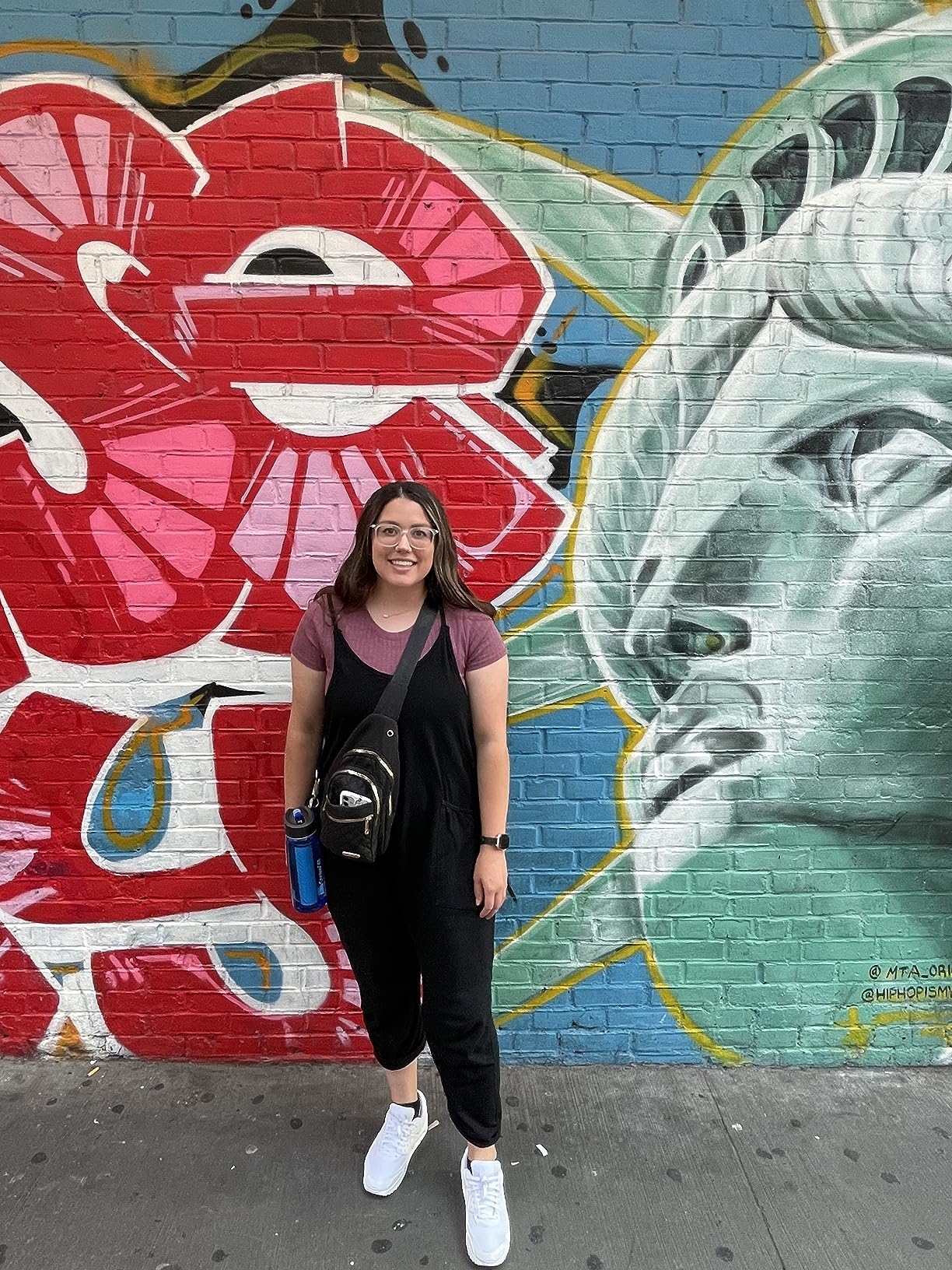 Person standing in front of a mural with the Statue of Liberty and flowers. They are wearing a red shirt and black overalls, holding a water bottle