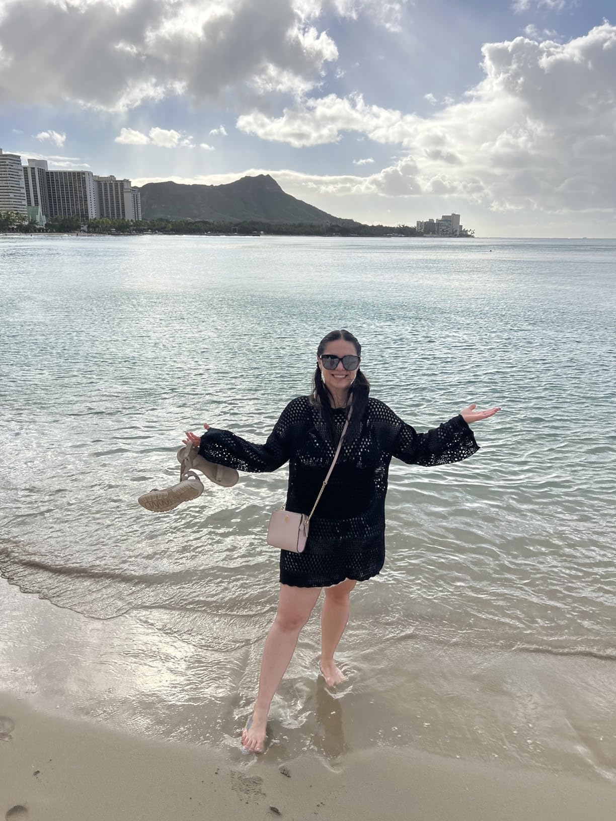 Reviewer on a beach holding sandals, wearing a black cover-up and sunglasses, smiling with Diamond Head and ocean in the background