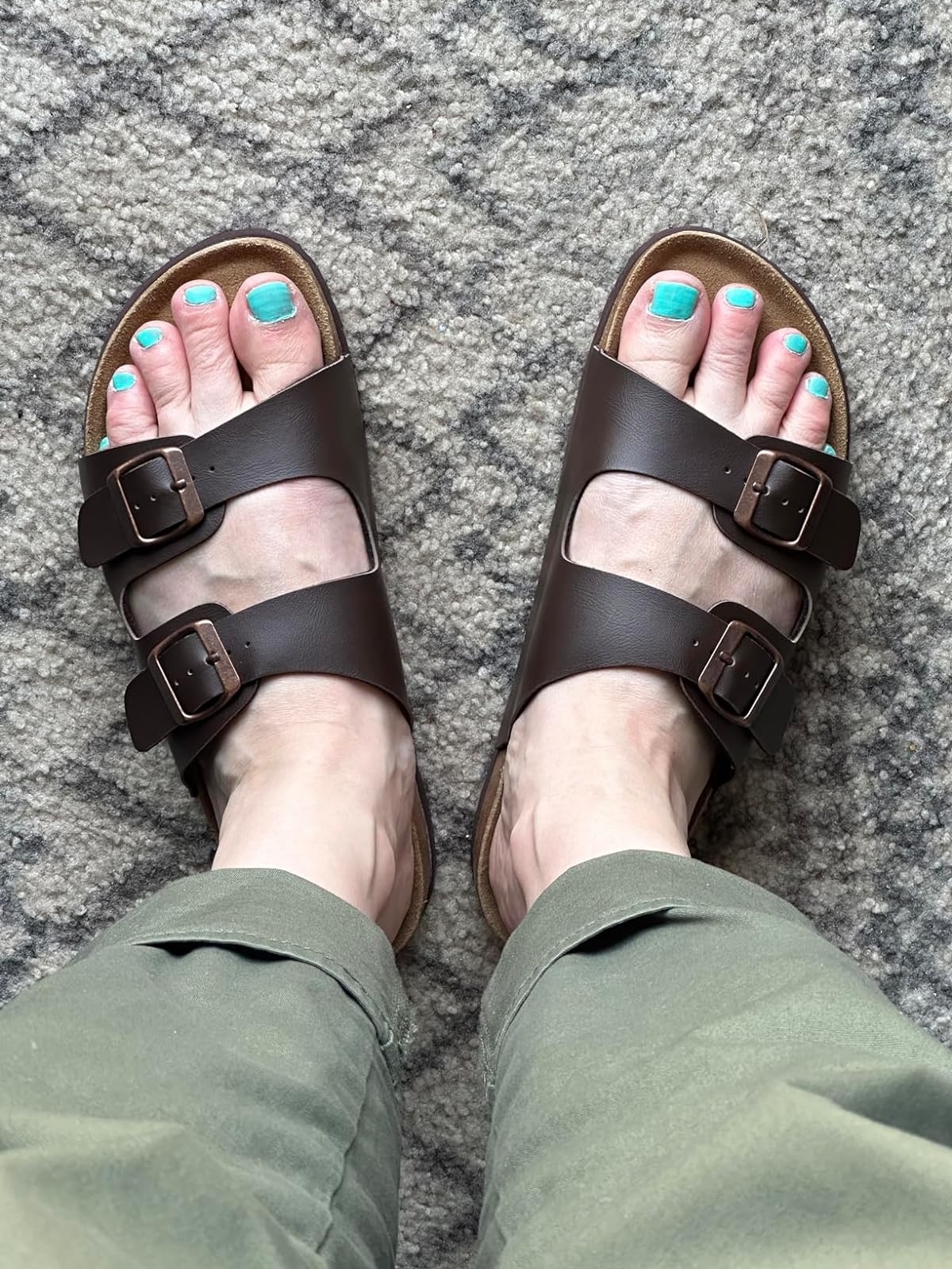 Person wearing brown two-strap sandals and green pants, with blue-painted toenails, standing on a textured surface