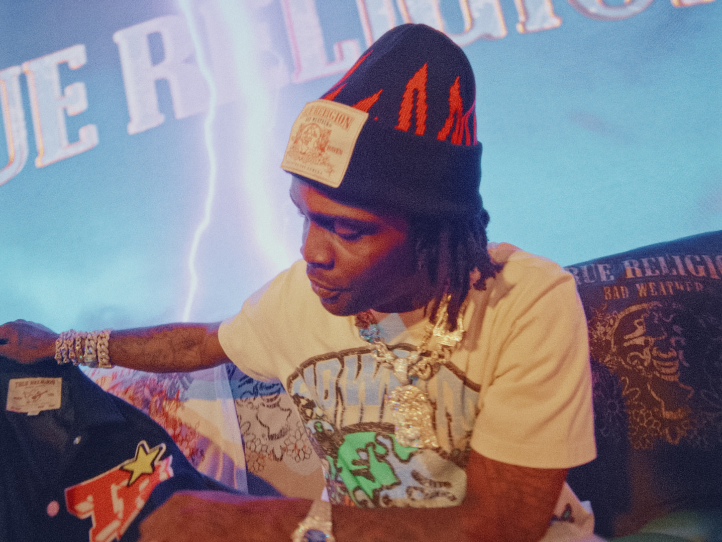 Man wearing a graphic t-shirt, black beanie, and multiple diamond chains, examining a jacket with patches, against a stylized background