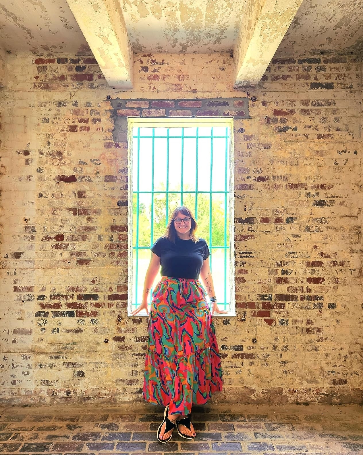 A reviewer stands in front of a window wearing a black top and a long, patterned skirt with flip-flops. The setting has rustic brick walls