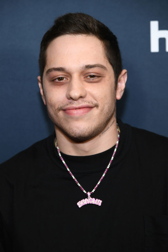 Pete Davidson smiling, wearing a black shirt and a necklace that says &quot;STENOLLI.&quot;