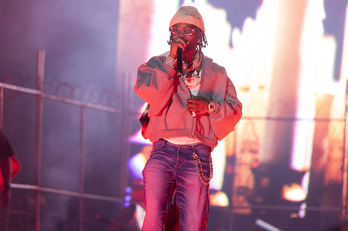 Offset performing on stage wearing a baggy sweatshirt, large chain necklaces, and sunglasses