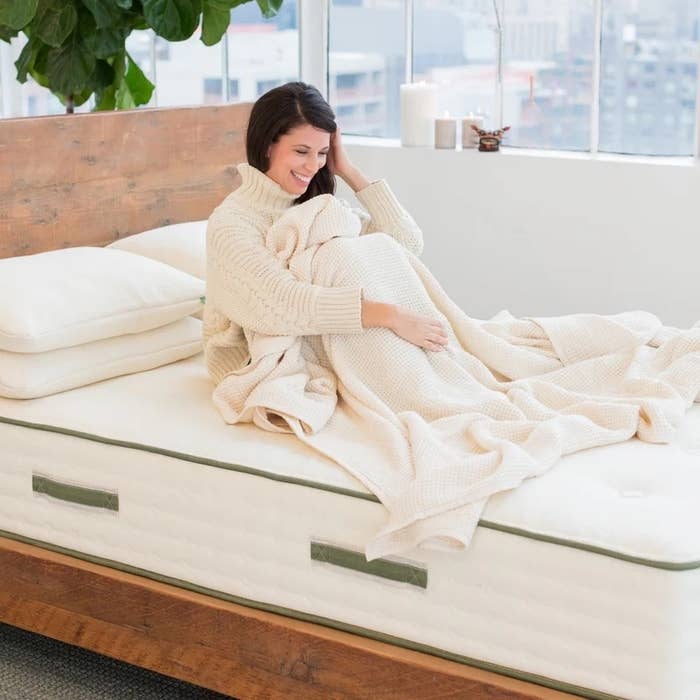 A woman in a cozy sweater sits on a neatly made bed in a modern, bright room, holding a blanket and smiling. This image accompanies a shopping article