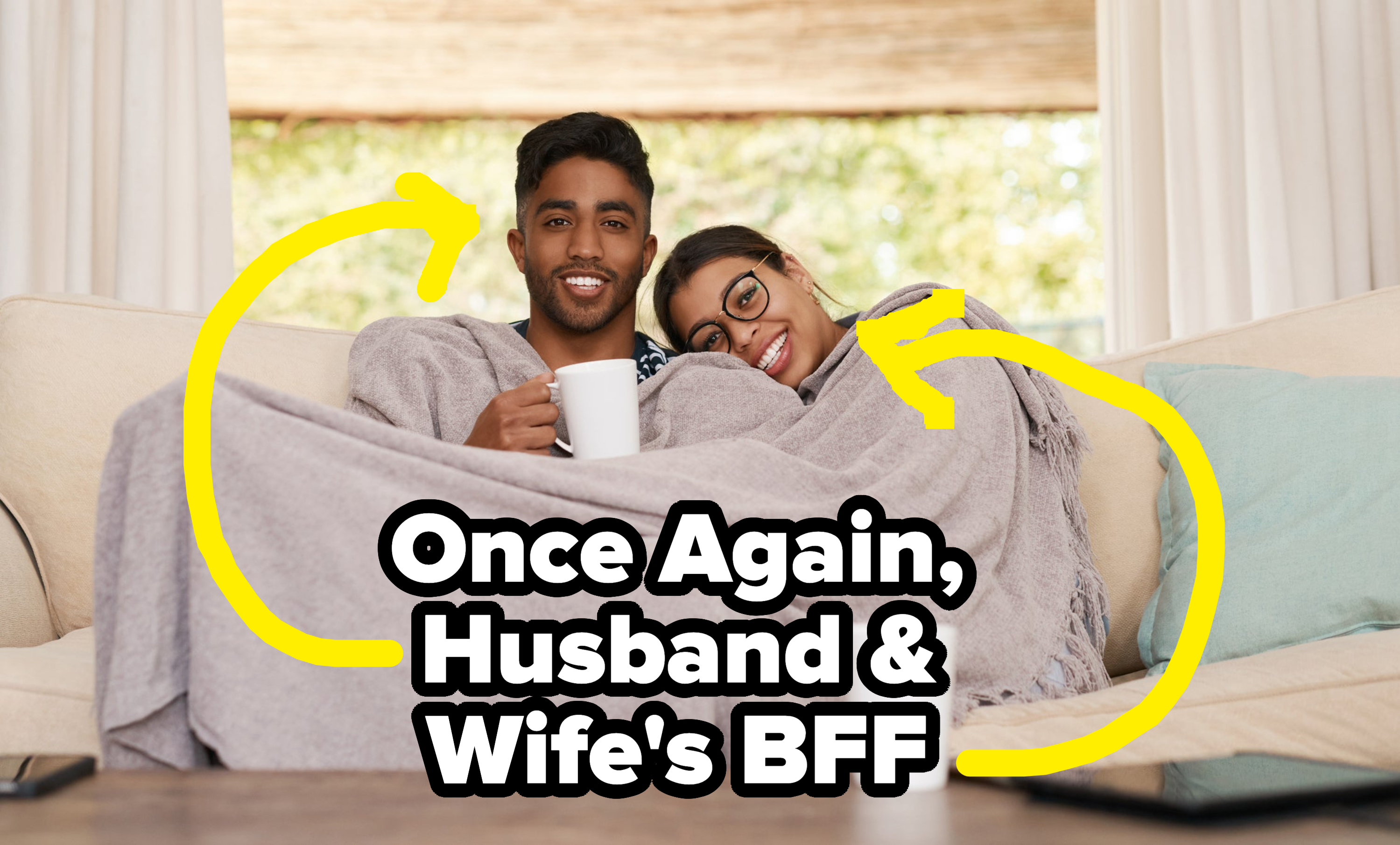 A couple snuggling under a blanket on a couch, smiling and holding mugs, surrounded by cozy home decor