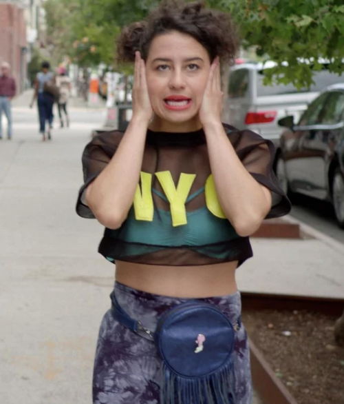 Ilana Glazer holds her face with both hands, wearing a black sheer crop top with &quot;NYC&quot; letters over a green tank and a blue floral skirt