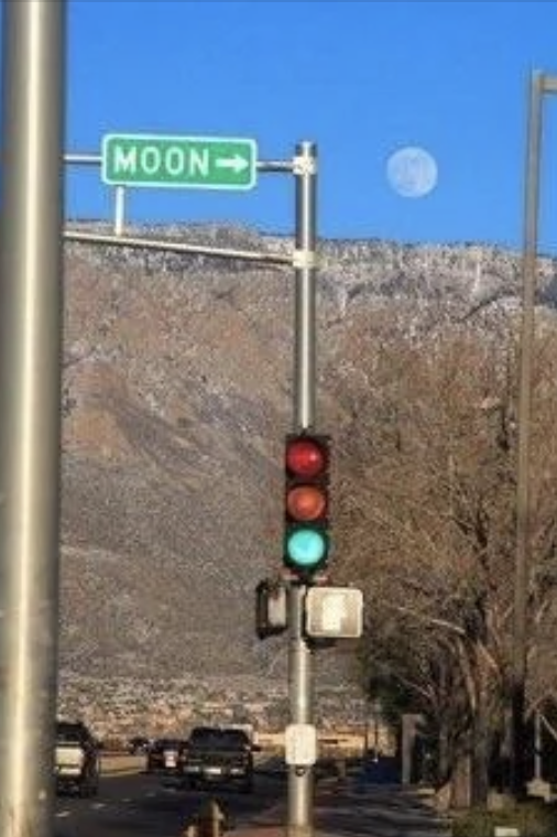A streetlight with a green light is under a green road sign that reads "MOON" with an arrow pointing right. The moon is visible in the sky