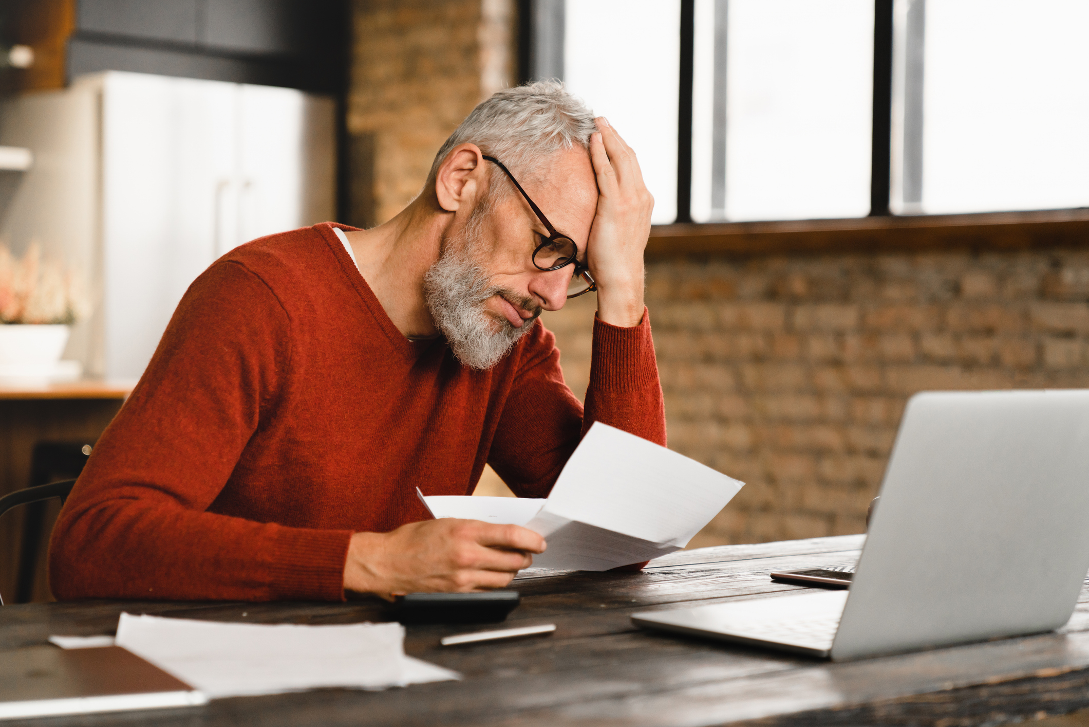 A man with gray hair and a beard wearing glasses is sitting at a desk with his hand on his head, looking stressed while reading a document, with a laptop nearby