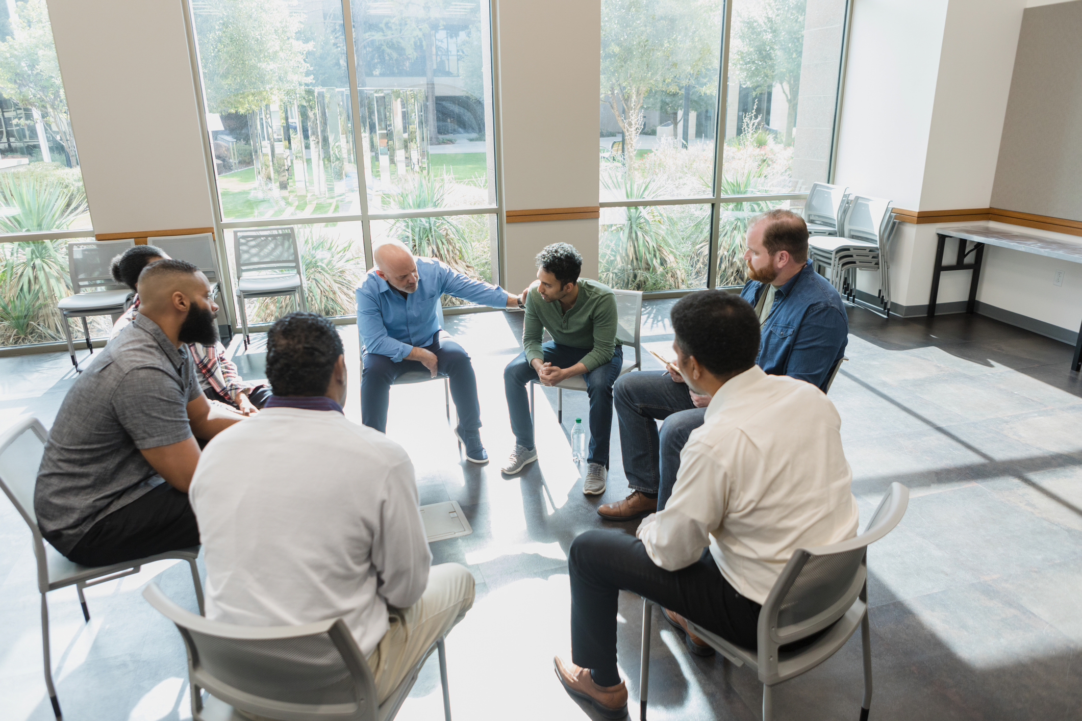 A group of seven men sit in a circle, engaged in a discussion in a brightly lit room with large windows