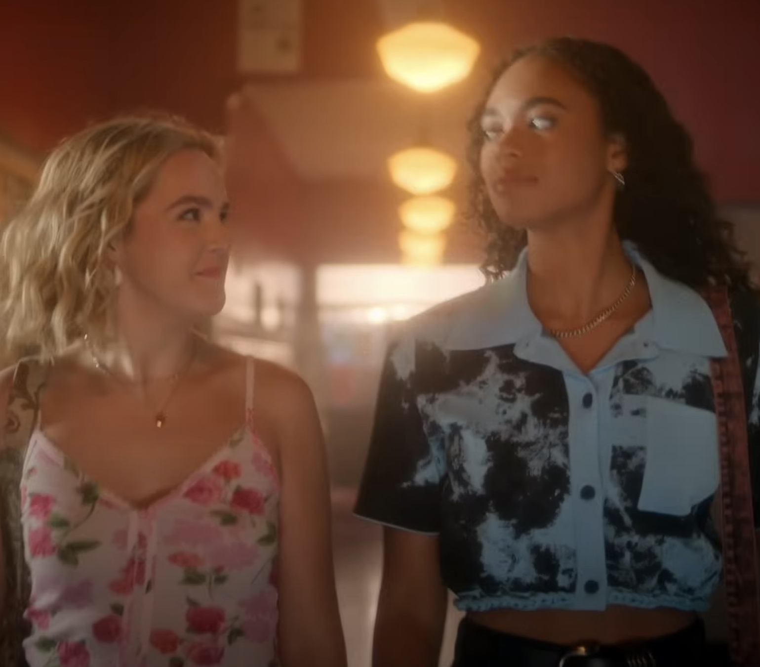 Elsie Fisher and Amiah Miller walk down a hallway. Elsie wears a floral tank top, and Amiah wears a tie-dye short-sleeved shirt with black pants