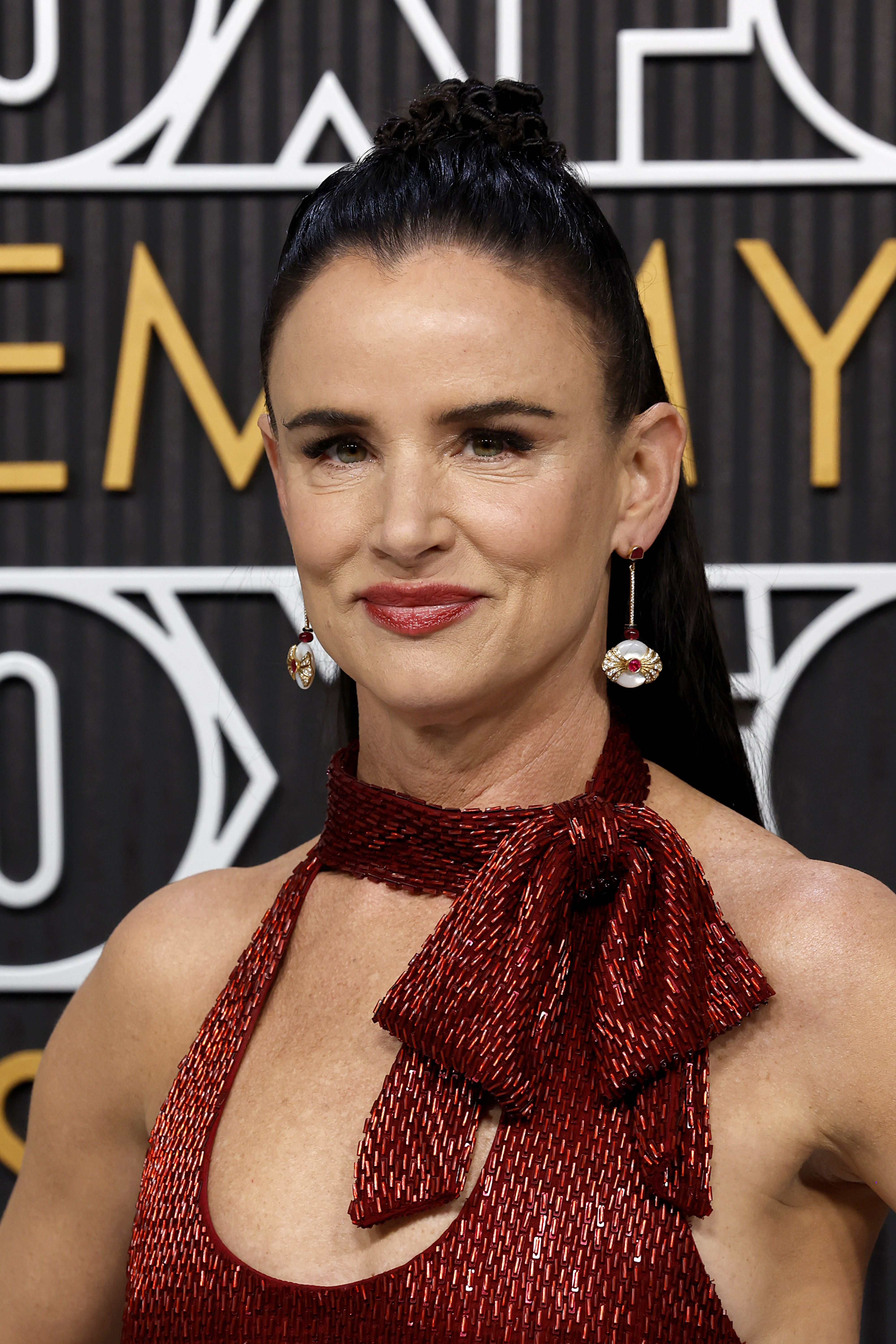 Juliette Lewis on the red carpet wearing a shimmering, sleeveless dress with a high neck bow detail and statement earrings