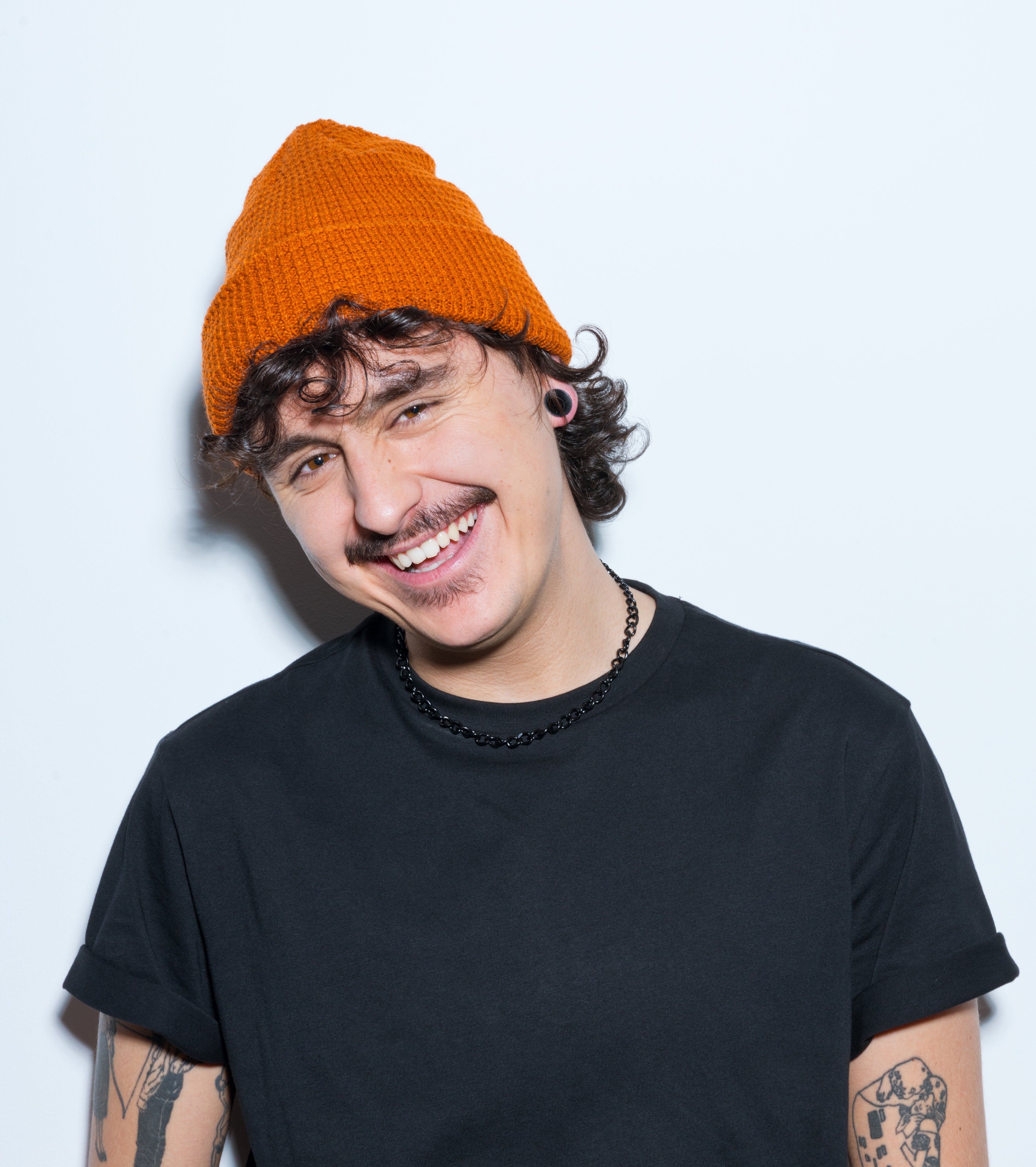 A person with curly hair wearing an orange beanie and a black T-shirt smiles at the camera. Tattoos are visible on their arms