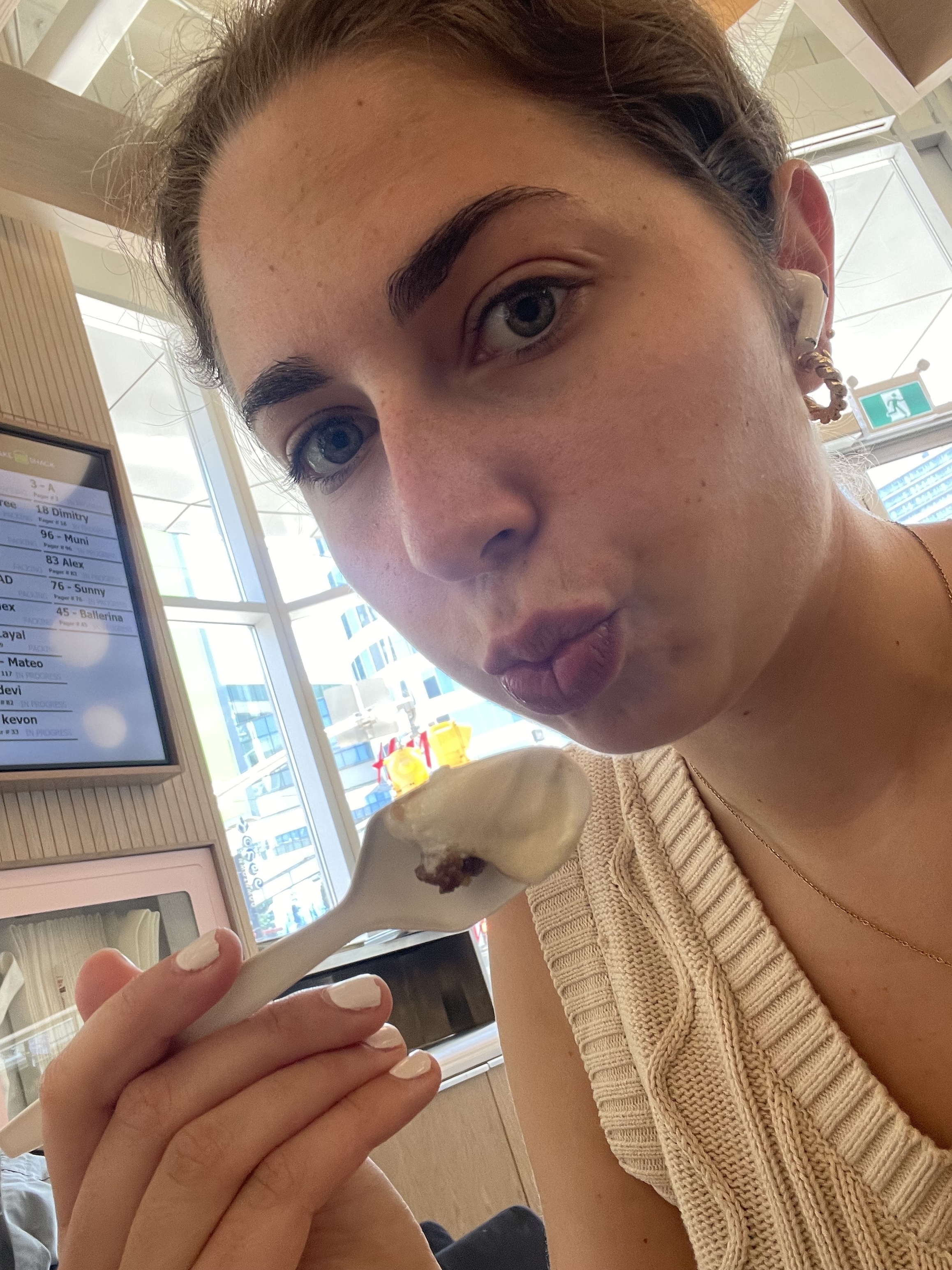 A woman in a sleeveless knit top holds a spoon with yogurt or ice cream near her mouth inside a modern café or restaurant with a digital menu in the background