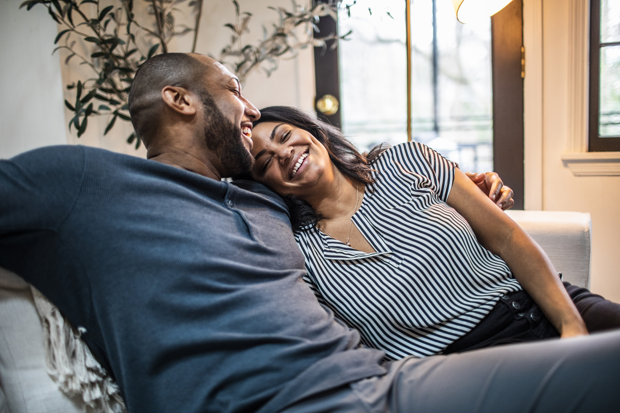 Man and woman smiling and cuddling on a sofa, wearing casual attire