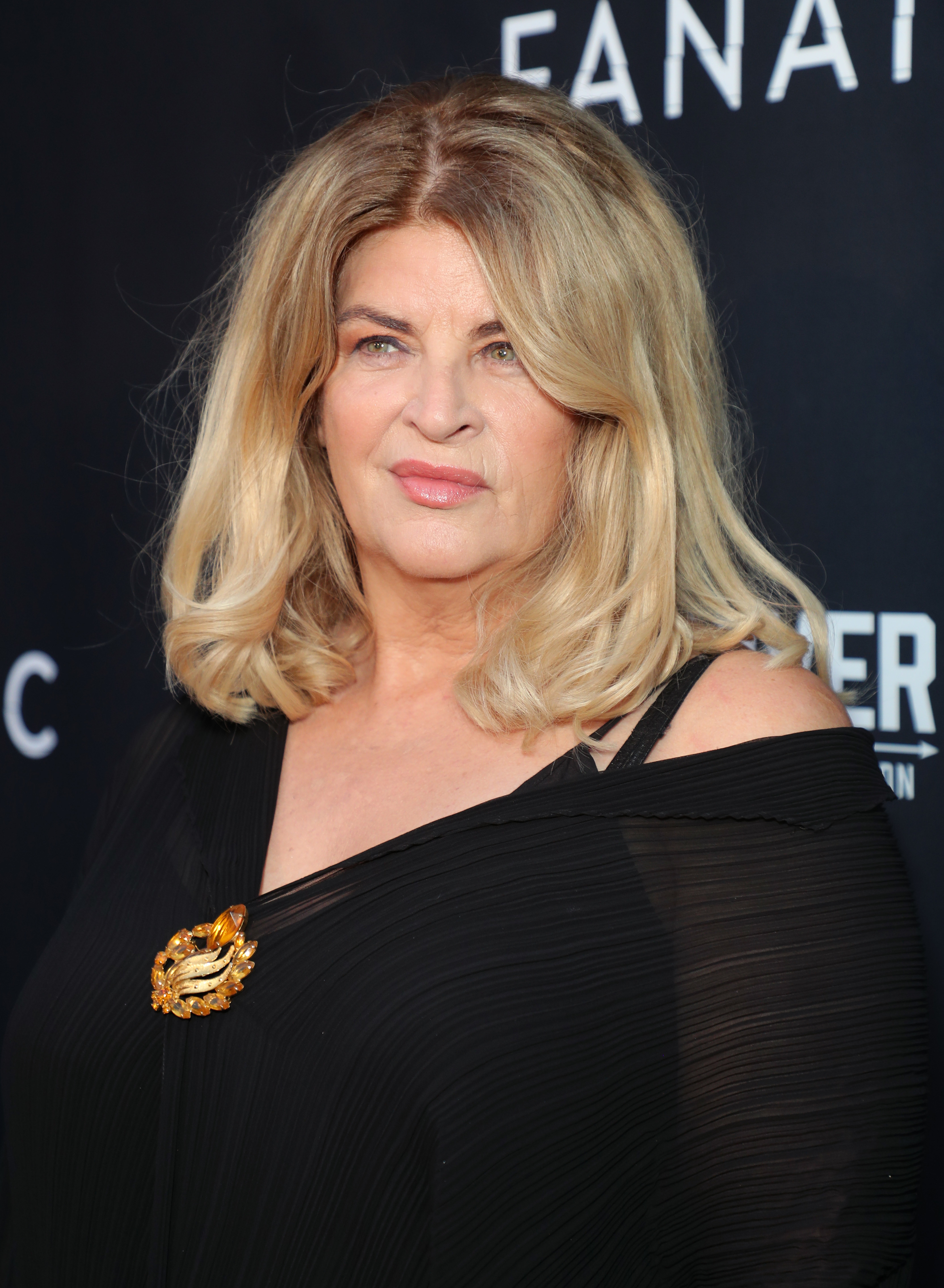 Kirstie Alley poses at a celebrity event, wearing an off-shoulder black dress with a decorative brooch