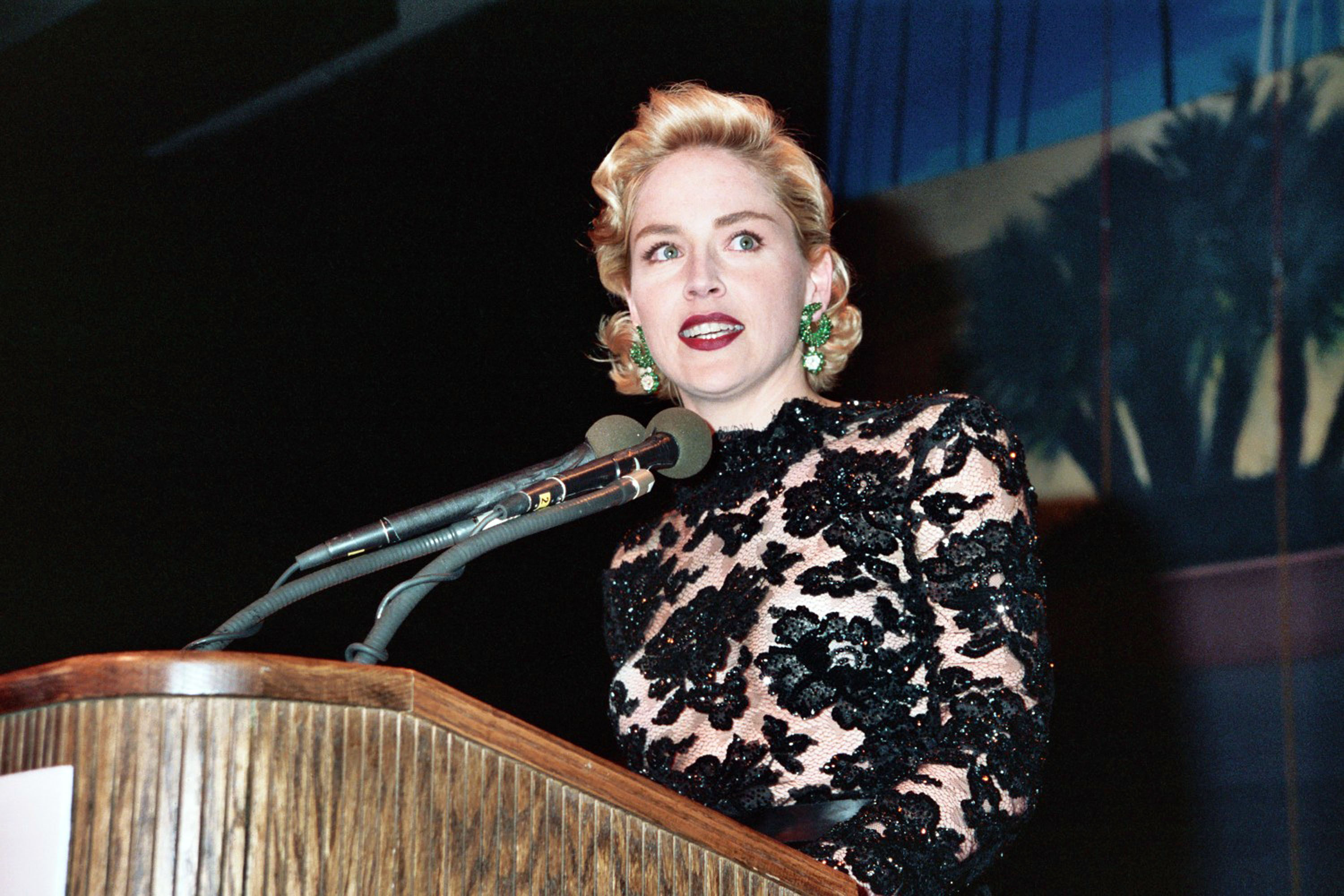 Sharon Stone, wearing a black lace top with bold green earrings, speaks at a podium during an event