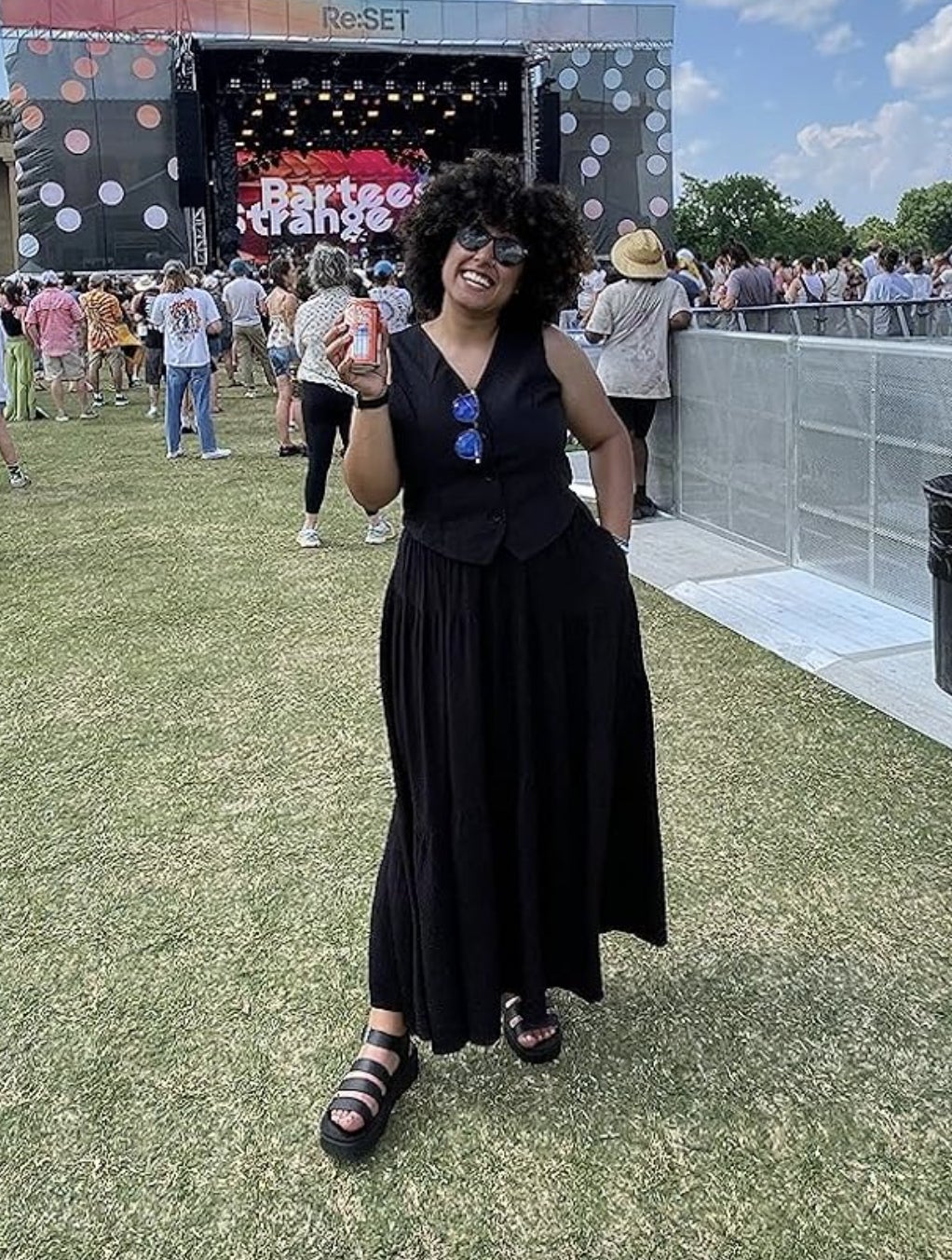 A idiosyncratic   wearing a sleeveless achromatic  dress, achromatic  vest, and sandals poses with a portion  tin  astatine  an outdoor euphony  festival. A signifier    and assemblage  are successful  the background