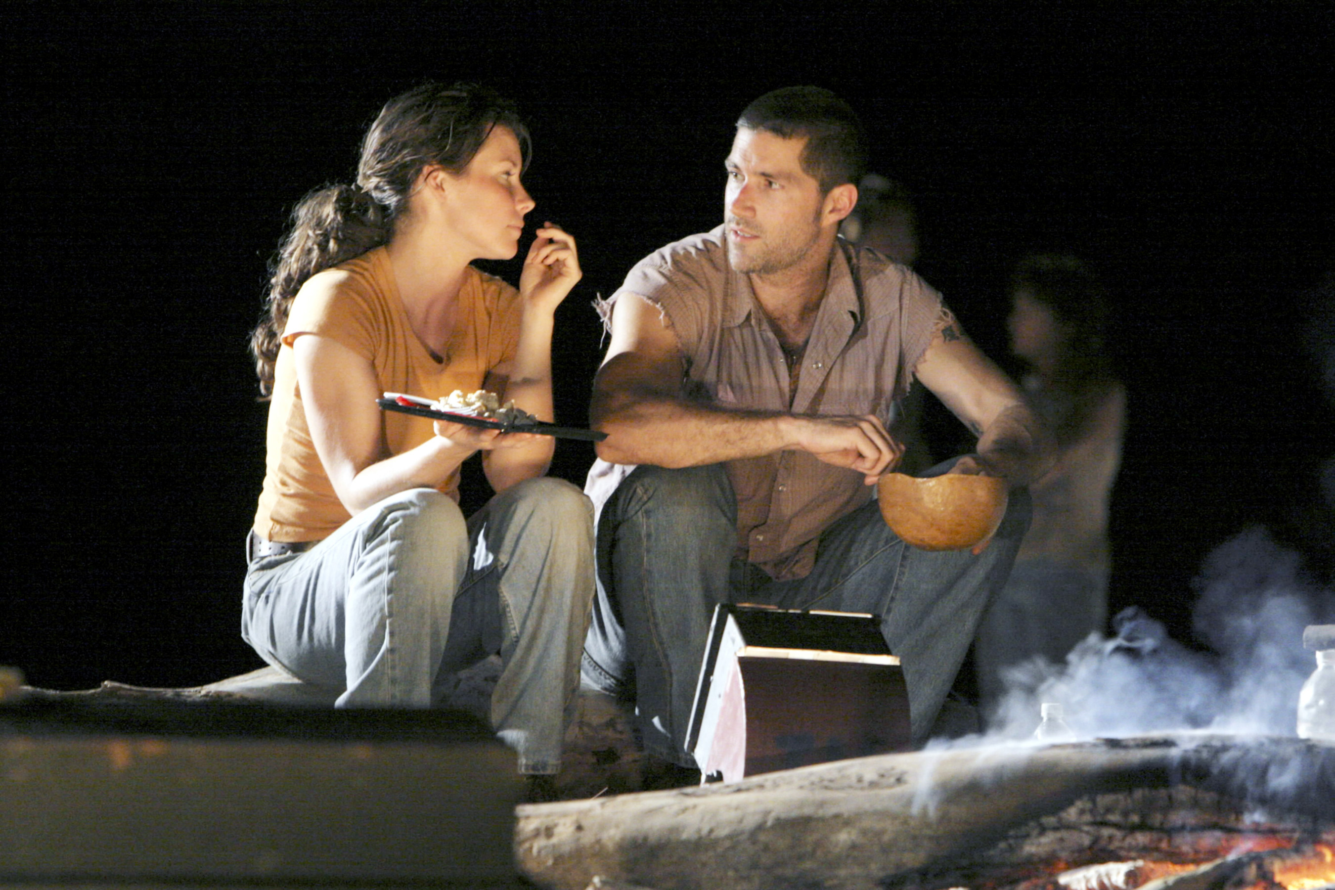 Evangeline Lilly and Matthew Fox sit by a campfire, eating and talking, possibly in a scene from a TV show or movie