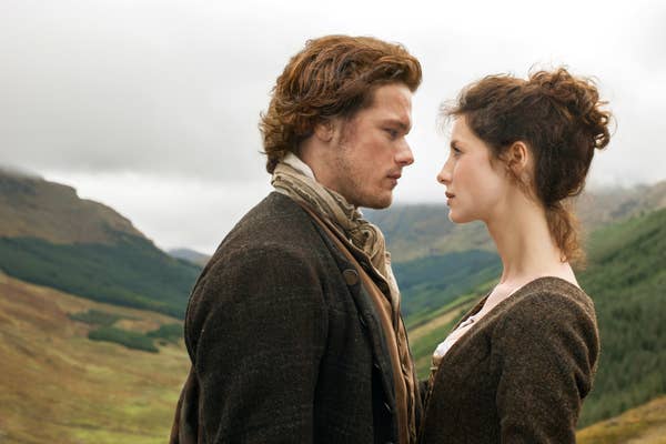 Sam Heughan and Caitríona Balfe in period clothing stand closely, gazing at each other in a scenic outdoor setting with hills in the background