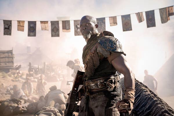 Djimon Hounsou stands in a post-apocalyptic setting holding a weapon, with fallen figures in the background, and flags hanging overhead