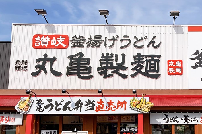 讃岐釜揚げうどんの店舗「丸亀製麺」の外観。うどん弁当直売所の看板と一緒に表示されています。