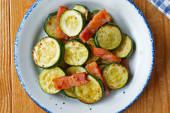 白い縁取りの皿に盛り付けられたズッキーニとベーコンの炒め物。チェック柄のナプキンとフォークが添えられています。