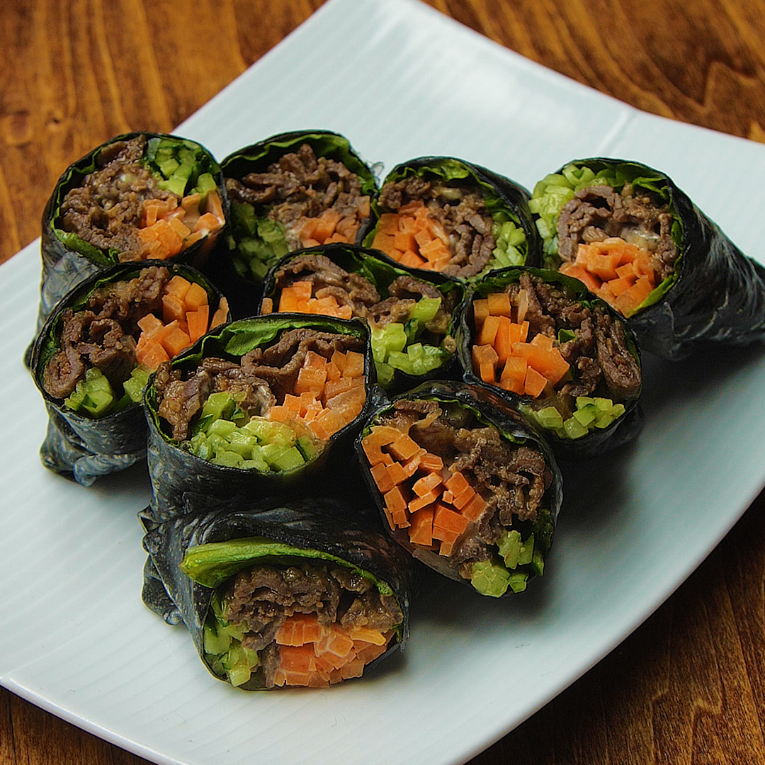 木製のテーブルの上に白い皿に盛られた牛肉、ニンジン、キュウリを巻いた海苔の巻きのりが並んでいます。