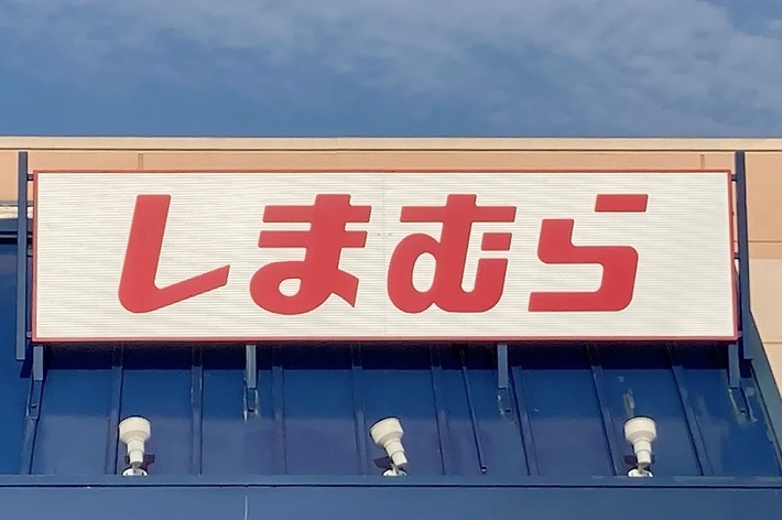 A storefront with a sign in Japanese text reading &ldquo;しまむら&rdquo; above the entrance. The blue awning below the sign shows part of a handicap parking sign