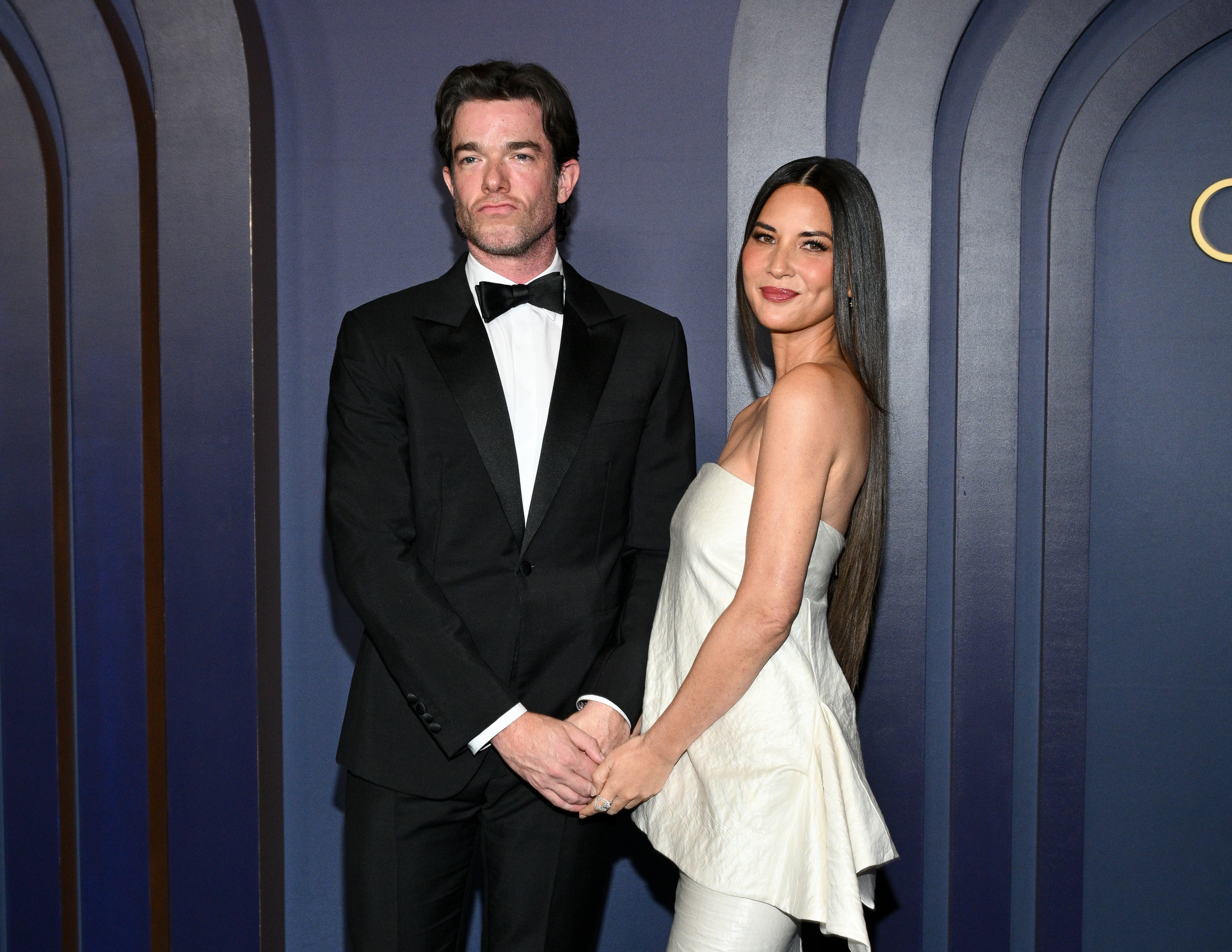 John Mulaney wears a tuxedo and Olivia Munn is in a strapless dress as they hold hands while posing for a photo at a formal event