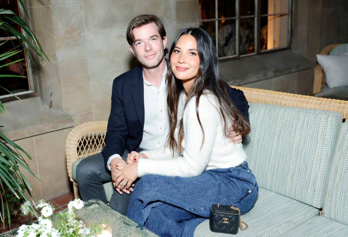 Olivia Munn and John Mulaney smiling, sitting close together on a bench, Olivia in a sweater and jeans, John in a blazer and shirt