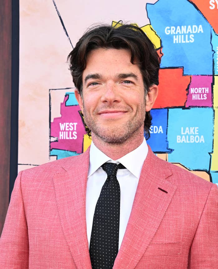 John Mulaney wearing a light suit and a polka dot tie in front of a colorful map backdrop