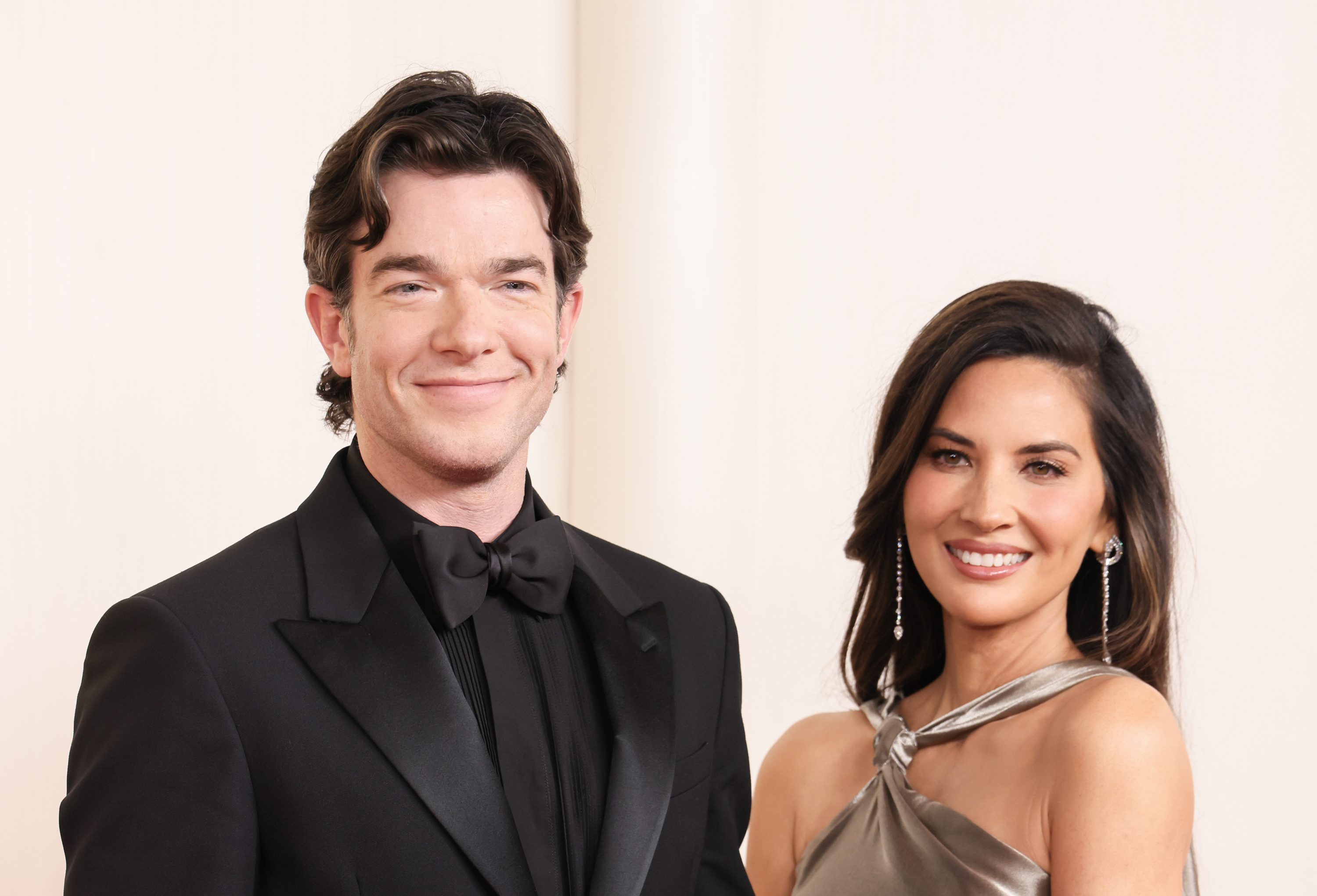 John Mulaney in a black tuxedo and Olivia Munn in a stylish one-shoulder dress smile while posing together