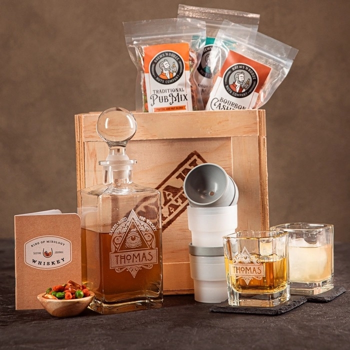 Personalized whiskey decanter and glasses set with a cocktail kit, pub mix, and cashews, displayed on a table next to a wooden crate