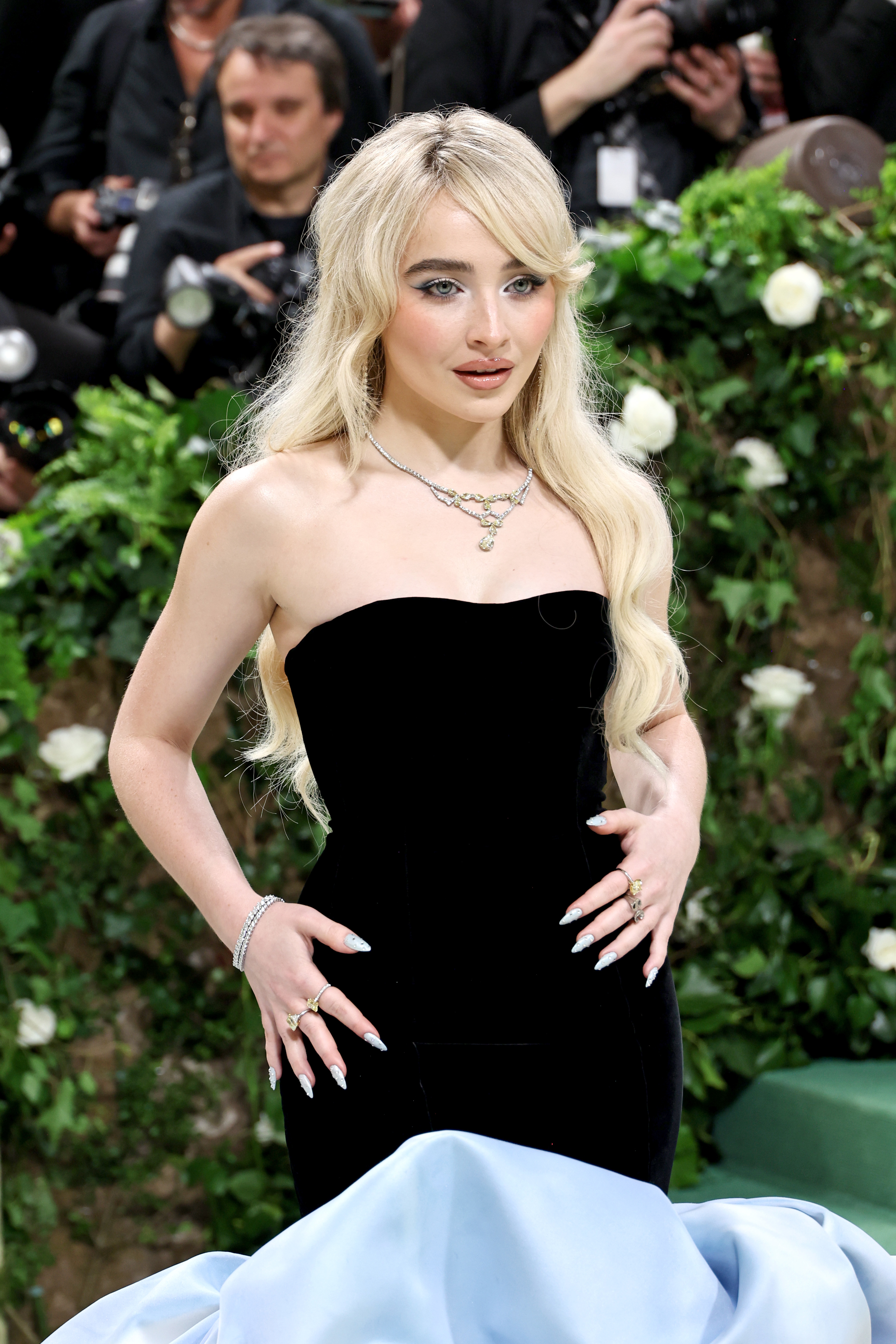 Sabrina Carpenter on a red carpet wearing a strapless, form-fitting dress with silver jewelry, posing in front of a floral backdrop