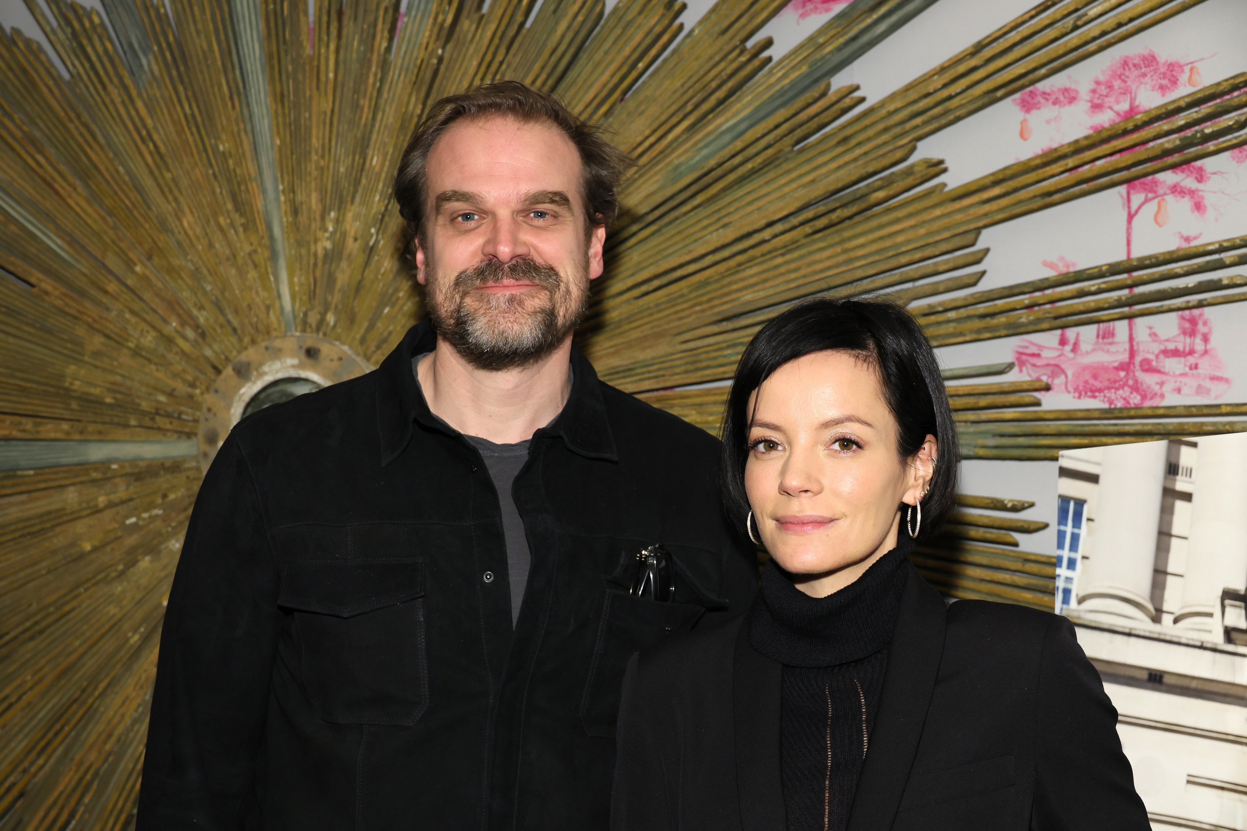 David Harbour and Lily Allen stand together, both wearing dark outfits, in front of a large artistic sunburst backdrop