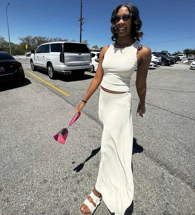 A reviewer is outdoors successful  a parking lot, wearing a sleeveless, agelong  achromatic  dress, sandals, and sunglasses, holding a pinkish  bag