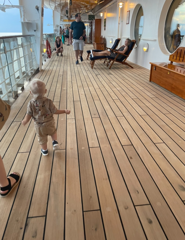 A toddler walks on the deck of a cruise ship, while adults lounge in chairs and others walk in the background