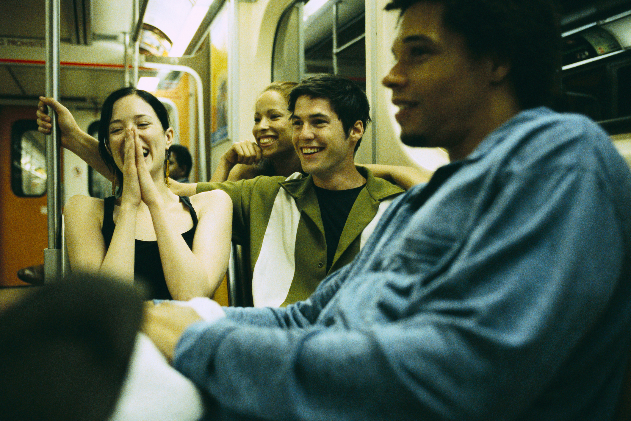 Four young adults, wearing casual clothing, laugh and chat while sitting on a train