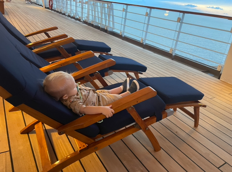 A child is lounging on a deck chair on a ship&#x27;s deck, with a calm sea visible in the background
