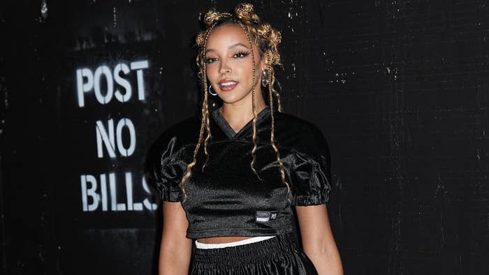 Singer Tinashe stands in front of a &quot;Post No Bills&quot; sign wearing a black cropped top with puff sleeves and matching pants, smiling for the camera